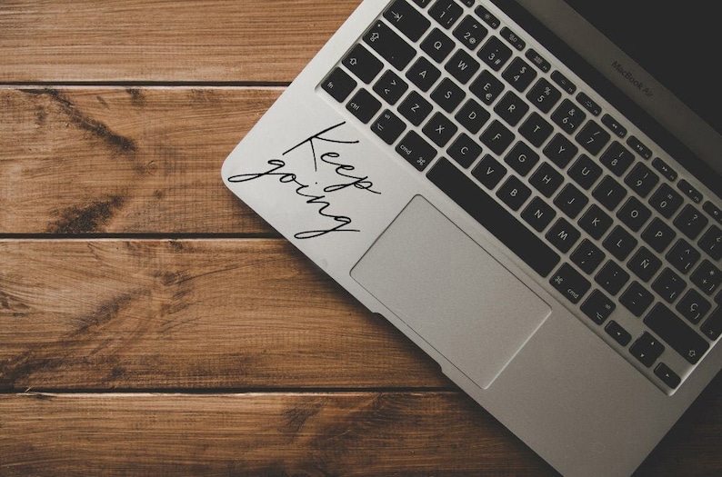 K&ouml;nnte beinhalten: Ein silberner Laptop mit einer schwarzen Tastatur und einem schwarzen Trackpad. Der Laptop hat einen Aufkleber mit der Aufschrift "Keep going".