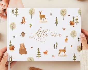 May include: A white baby memory book with a woodland theme. The cover features a gold inscription that reads "Little One" and is decorated with illustrations of animals like deer, bears, raccoons, and squirrels.