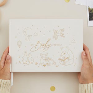 May include: A cream-coloured baby journal with gold foil illustrations of a moon, hot air balloon, stork, ducks, deer, and a bear reading. The words "My Baby Journal" are written in gold script. The journal is held by a person wearing a cream-coloured sweater.