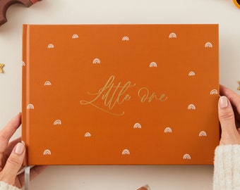 Könnte beinhalten: Ein braunes Hardcover-Fotoalbum mit einem weißen Band. Der Einband hat eine goldene Aufschrift mit der Aufschrift "Little One" und ist mit einem Muster aus weißen Regenbögen verziert.
