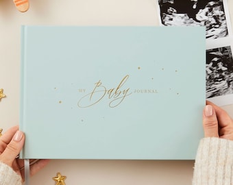 May include: A light blue baby journal with gold lettering that reads "My Baby Journal". The journal is decorated with gold stars and is being held by a person's hands.