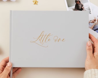 May include: A gray photo album with the words "Little One" in gold lettering. The album is being held by a person's hands. The background is a light beige color with white clouds and gold stars.