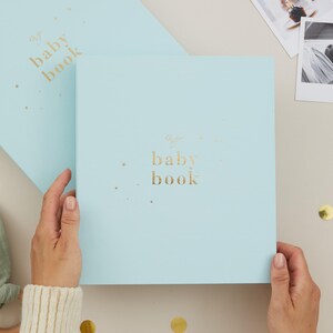 May include: A light blue baby book with gold lettering that reads "my baby book". The book is square-shaped and has a matching light blue cover. The image also includes a baby rattle and photos.
