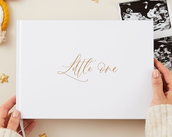 Könnte beinhalten: Weißes Baby-Erinnerungsbuch mit goldener Aufschrift "Little One". Das Buch liegt auf einer hellbeigen Oberfläche mit goldenen Sternen und einem regenbogenfarbenen Babyspielzeug.