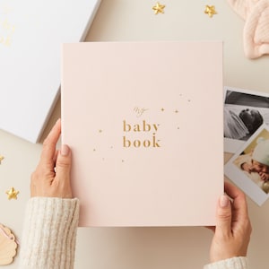 May include: A pink baby book with gold foil lettering that reads "my baby book" and gold stars scattered across the cover.