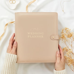 Könnte beinhalten: Ein beigefarbener Hochzeitsplaner mit den Worten "Our Wedding Planner" auf dem Einband. Der Planer wird von einer Person gehalten, die einen cremefarbenen Pullover trägt. Goldene Eheringe und eine Halskette sind im Hintergrund.