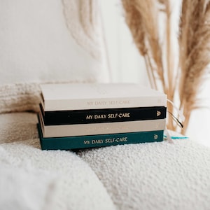 Op de afbeelding: Een stapel van vier dagboeken met de tekst "My Daily Self-Care" op de voorkant. De dagboeken zijn in verschillende kleuren: cr&egrave;me, zwart, cr&egrave;me en teal.