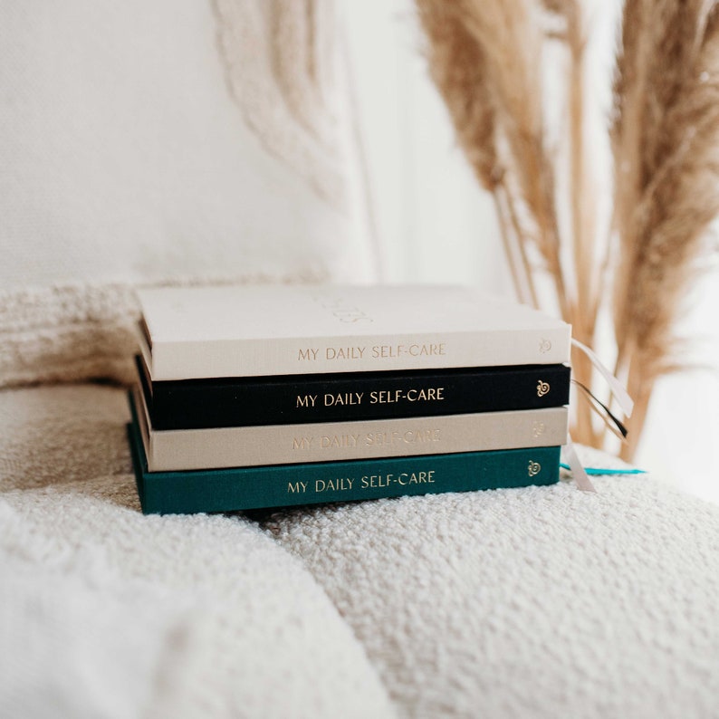Op de afbeelding: Een stapel van vier dagboeken met de tekst "My Daily Self-Care" op de voorkant. De dagboeken zijn in verschillende kleuren: cr&egrave;me, zwart, cr&egrave;me en teal.