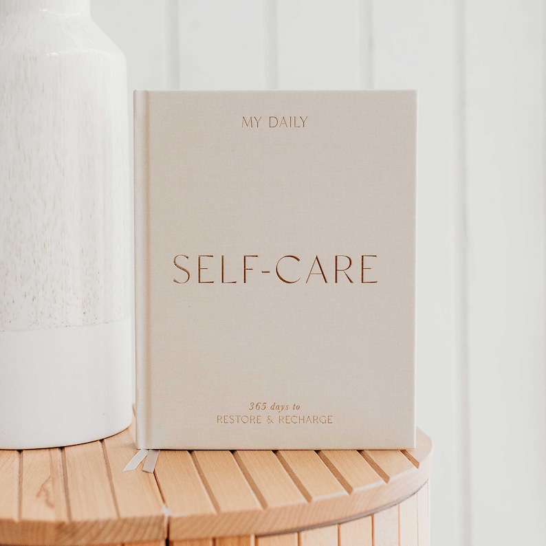Op de afbeelding: Een cr&egrave;mekleurig dagboek met de tekst "MY DAILY SELF-CARE 365 days to RESTORE & RECHARGE" op de voorkant.