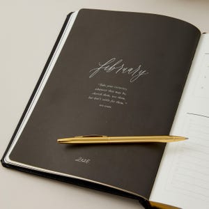 Peut inclure: Journal noir ouvert avec le mot "february" en &eacute;criture cursive. Un stylo dor&eacute; repose sur la page avec une citation de Mia Hamm. L'ann&eacute;e "2026" est imprim&eacute;e au bas de la page.
