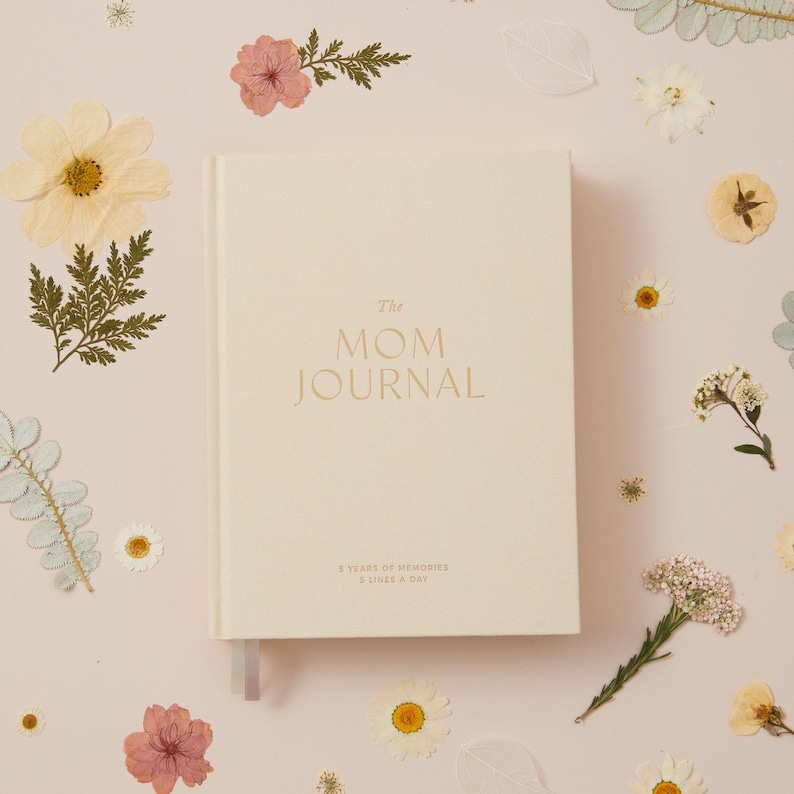 May include: Cream-colored journal titled "The Mom Journal" with gold lettering. The journal is surrounded by dried flowers and greenery on a light pink background. The journal includes a ribbon bookmark.
