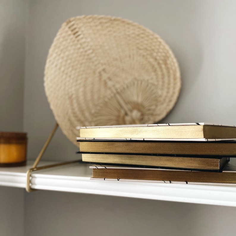 May include: A stack of three hardcover books with gold-gilded edges and black spines rests on a white shelf. A woven fan and a small candle sit in the background. The books have a minimalist design.
