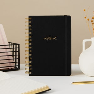 May include: A black spiral bound notebook with a gold coil and a gold inscription that reads "notebook".