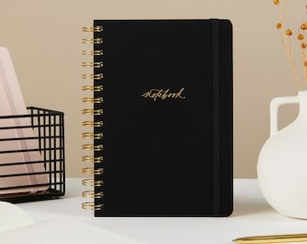 May include: A black spiral bound notebook with a gold coil and a gold inscription that reads "notebook".