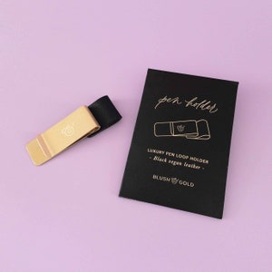 May include: A gold pen loop holder with a black vegan leather strap. The holder features a small engraved ampersand symbol. The packaging is black with gold lettering and says "pen holder" and "Luxury Pen Loop Holder - Black vegan leather - Blush & Gold".