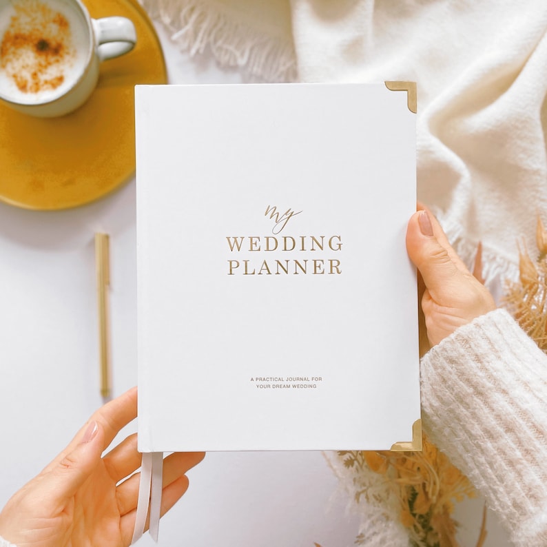 K&ouml;nnte beinhalten: Ein wei&szlig;er Hochzeitsplaner mit goldener Beschriftung, die "my wedding planner" und "a practical journal for your dream wedding" lautet.