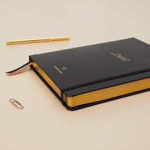 Peut inclure: Un agenda noir de 2026 avec des bords dor&eacute;s et un marque-page en ruban assorti. Un stylo et un trombone dor&eacute;s sont &eacute;galement pr&eacute;sents. L'agenda porte le texte "Week by Week" et un logo sur la couverture.