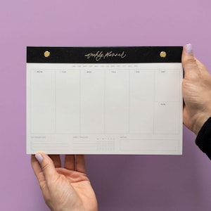 Könnte beinhalten: Ein schwarz-weißer Wochenplaner mit goldenen Akzenten. Auf der Oberseite des Planers steht in goldener Schrift "weekly planner". Der Planer wird vor einem lila Hintergrund gehalten. Er hat Abschnitte für jeden Wochentag, Top-Prioritäten, einen Gewohnheitstracker und Notizen.