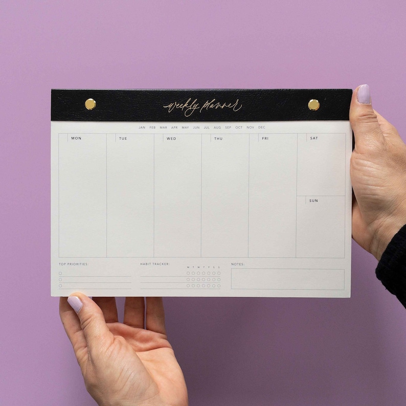 K&ouml;nnte beinhalten: Ein schwarz-wei&szlig;er Wochenplaner mit goldenen Akzenten. Auf der Oberseite des Planers steht in goldener Schrift "weekly planner". Der Planer wird vor einem lila Hintergrund gehalten. Er hat Abschnitte f&uuml;r jeden Wochentag, Top-Priorit&auml;ten, einen Gewohnheitstracker und Notizen.