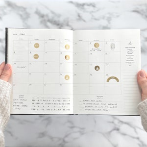 May include: Open planner showing the month of May with gold foil stickers marking dates. The planner includes sections for habits and monthly goals. The text "Dinner with Sue" is visible on the 5th, and "Holiday!" is written on the 14th.