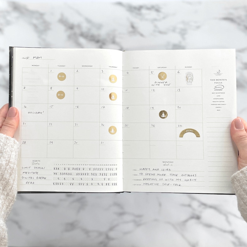 May include: Open planner showing the month of May with gold foil stickers marking dates. The planner includes sections for habits and monthly goals. The text "Dinner with Sue" is visible on the 5th, and "Holiday!" is written on the 14th.