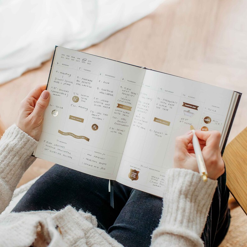 May include: A black and gold planner open to a weekly spread, with handwritten notes and gold foil accents. A person is writing in the planner with a gold pen. The planner is open on a person's lap.