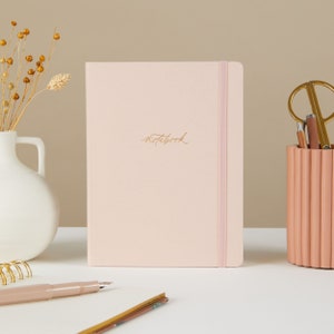 Peut inclure: Un carnet rose avec une fermeture élastique dorée et le mot "notebook" écrit en lettres dorées sur la couverture.
