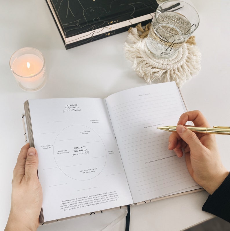May include: An open journal with the text "Let go of the things you cannot control" and "Focus on the things you can control." A gold pen is held over the lined pages. A lit candle, a glass of water, and a macrame coaster are also visible.