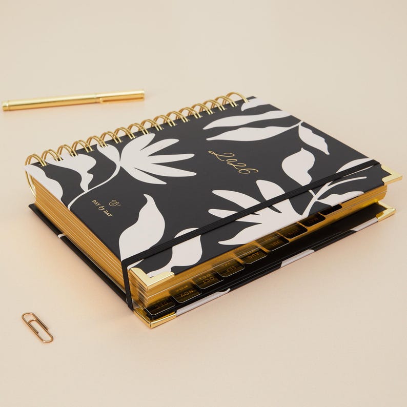 May include: A black and white floral patterned planner with gold accents. The planner has a gold spiral binding, gold-edged pages, and a black elastic band. The year "2026" is printed in gold on the cover. A gold pen and paperclip are also visible.