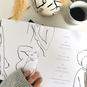 May include: Open planner with line art illustrations and chapter headings: Intentions, Big Picture, Week by Week, and Reflection Journal. A cup of coffee and a decorative vase are in the background.