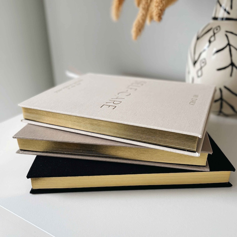 Op de afbeelding: Drie hardcover-dagboeken gestapeld op elkaar. Het bovenste dagboek is beige met gouden randen en de tekst "SELF-CARE" in reli&euml;f op de omslag. Het middelste dagboek is grijs met gouden randen. Het onderste dagboek is zwart met gouden randen.