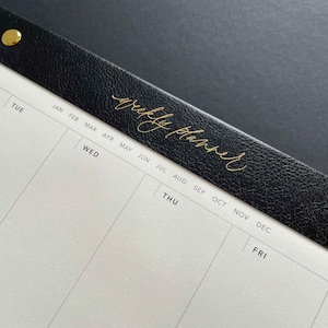 K&ouml;nnte beinhalten: Nahaufnahme eines Wochenplaners. Der schwarze Ledereinband ist mit den Worten "weekly planner" in goldener Schrift gepr&auml;gt. Die Seiten des Planers sind wei&szlig; mit grauen Linien, und die Wochentage und Monate sind in Schwarz gedruckt.