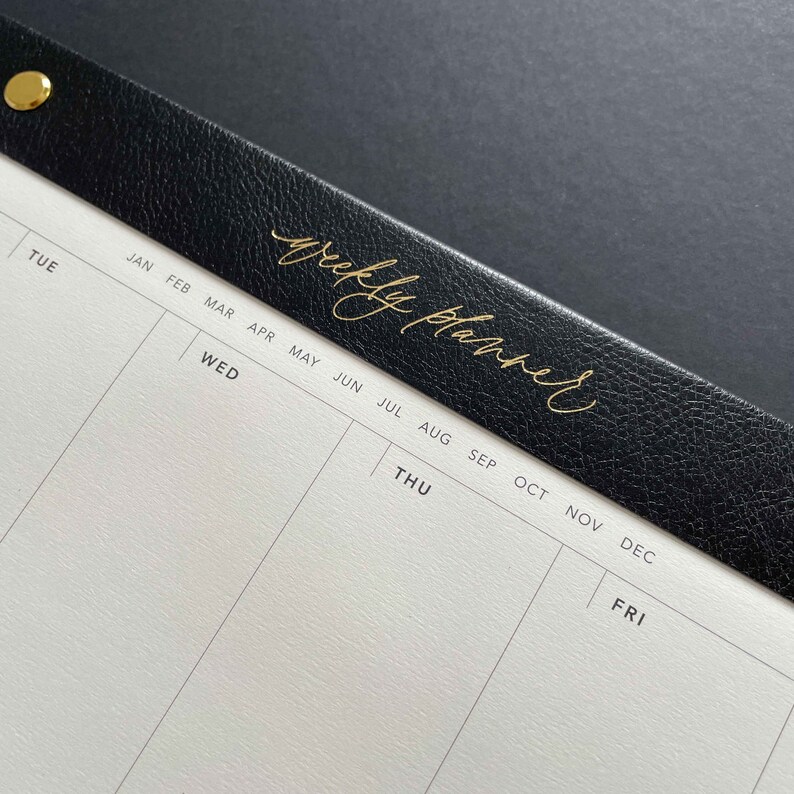 K&ouml;nnte beinhalten: Nahaufnahme eines Wochenplaners. Der schwarze Ledereinband ist mit den Worten "weekly planner" in goldener Schrift gepr&auml;gt. Die Seiten des Planers sind wei&szlig; mit grauen Linien, und die Wochentage und Monate sind in Schwarz gedruckt.
