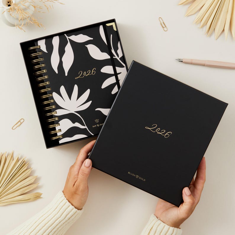 May include: A black 2026 planner with a floral design and gold spiral binding, presented in a matching box. The planner features a black cover with white floral patterns and the year 2026 in gold script.