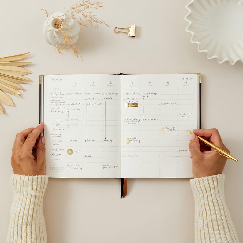 Peut inclure: Un agenda noir ouvert avec des d&eacute;tails dor&eacute;s, pr&eacute;sentant un calendrier mensuel. Un stylo dor&eacute; est utilis&eacute; pour &eacute;crire dans l'agenda. Des &eacute;l&eacute;ments d&eacute;coratifs incluent un vase blanc, une pince &agrave; reliure dor&eacute;e et des plantes s&eacute;ch&eacute;es. L'agenda est ouvert sur la page de f&eacute;vrier.