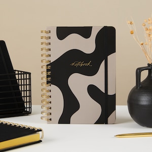 Puede incluir: Un cuaderno con espiral y un diseño abstracto en negro y beige. La portada presenta la palabra "notebook" en letras doradas. El cuaderno tiene una encuadernación en espiral dorada y un cierre elástico negro. Una cesta de alambre negra y un jarrón negro están en el fondo.