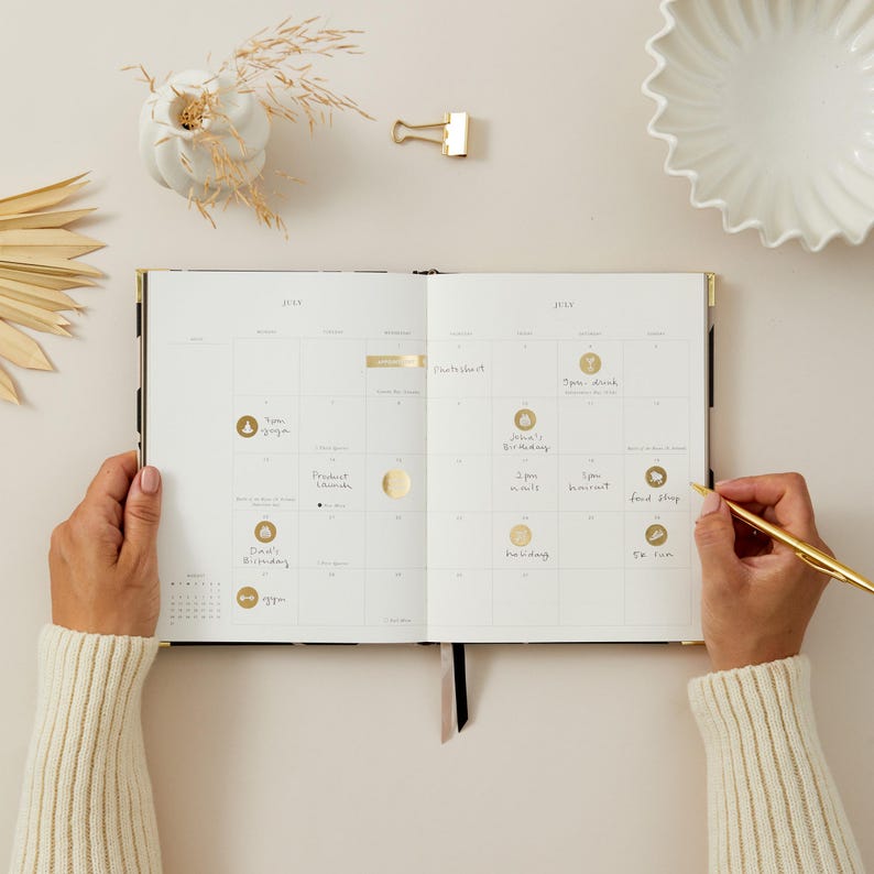 May include: An open planner with a July calendar spread, featuring gold accents and handwritten notes. A gold pen is held in the right hand, while the left hand holds the planner open. The planner is on a neutral surface with decorative items.