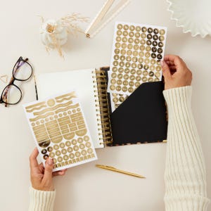 May include: A gold spiral-bound planner with a black pocket and gold stickers. The stickers are gold and white with various shapes and icons. A gold pen and glasses are also on the white surface.