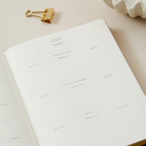 Peut inclure: Un carnet ouvert avec le titre "2026 Goals" et des sections pour la carri&egrave;re, le d&eacute;veloppement personnel et les voyages. Un trombone dor&eacute; est pos&eacute; sur la page. Le carnet est ouvert sur une page avec des invites pour les objectifs &agrave; court, moyen et long terme.