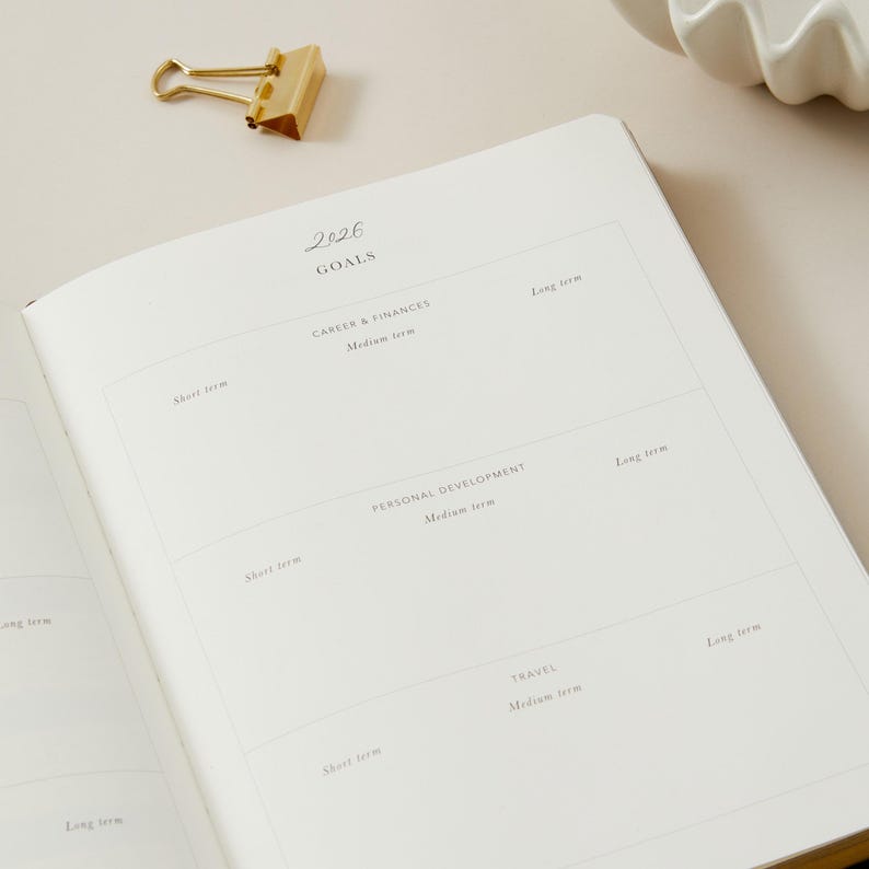 Peut inclure: Un carnet ouvert avec le titre "2026 Goals" et des sections pour la carri&egrave;re, le d&eacute;veloppement personnel et les voyages. Un trombone dor&eacute; est pos&eacute; sur la page. Le carnet est ouvert sur une page avec des invites pour les objectifs &agrave; court, moyen et long terme.