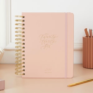 May include: A blush-colored spiral-bound planner with gold lettering that reads "Twenty Twenty Six". The planner has a matching elastic band closure and gold spiral binding. A pen holder with pens and a gold pen are visible.