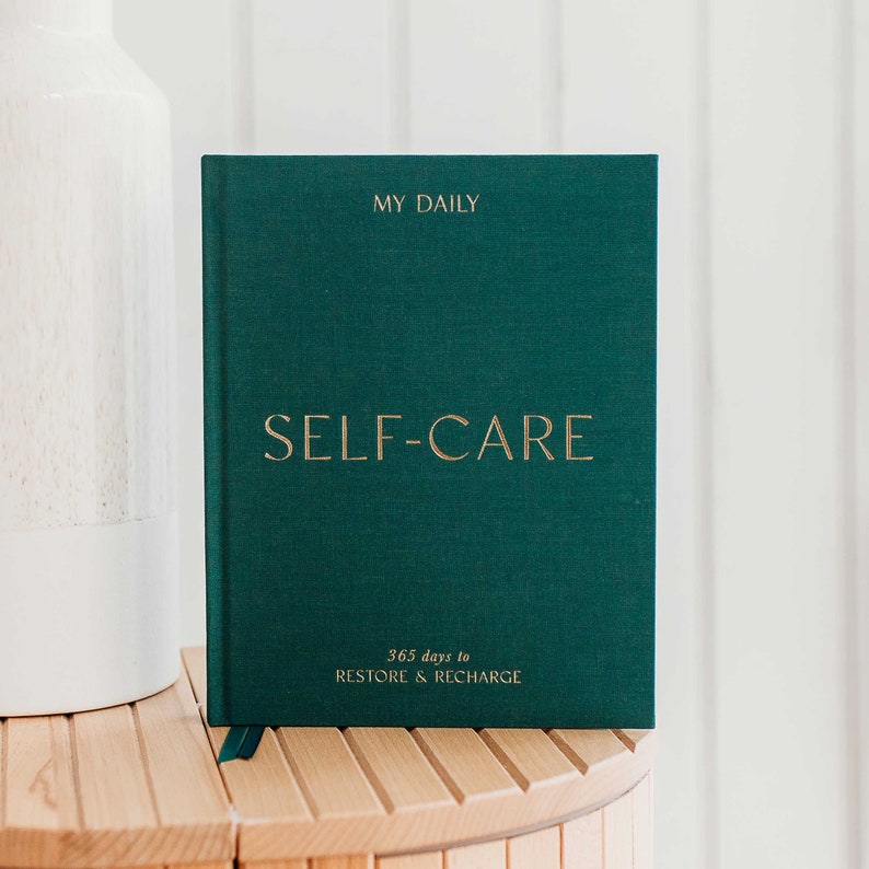 Puede incluir: Un diario de tapa dura verde con letras doradas que dicen "My Daily Self-Care 365 days to Restore & Recharge".