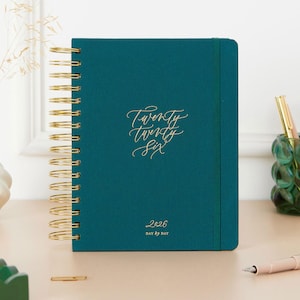 Könnte beinhalten: Türkisfarbener 2026-Planer mit goldener Spiralbindung. Das Cover zeigt goldfarbenen Schriftzug "Twenty Twenty Six" und "Day by Day". Ein goldener Stift und ein grüner Glas-Stifthalter sind sichtbar.