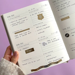 May include: A close-up of a daily planner with gold accents and stickers. The planner has a daily gratitude section with handwritten entries. The planner is open to pages 209, 210, and 212. The planner has a gold sticker that says "You can't pour from an empty cup".