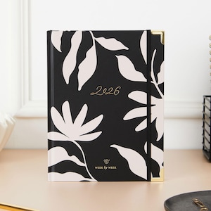 May include: A black and white floral 2026 planner with gold accents. The cover features a repeating pattern of white leaves and flowers against a black background. The year "2026" is written in gold script. The planner has gold corner protectors and the text "Week by Week".