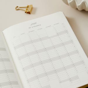 Peut inclure: Agenda ouvert avec un calendrier 2026, pr&eacute;sentant des grilles mensuelles et des dates. Un trombone dor&eacute; est pos&eacute; en haut. Les pages de l'agenda sont claires, avec un design simple et &eacute;l&eacute;gant.