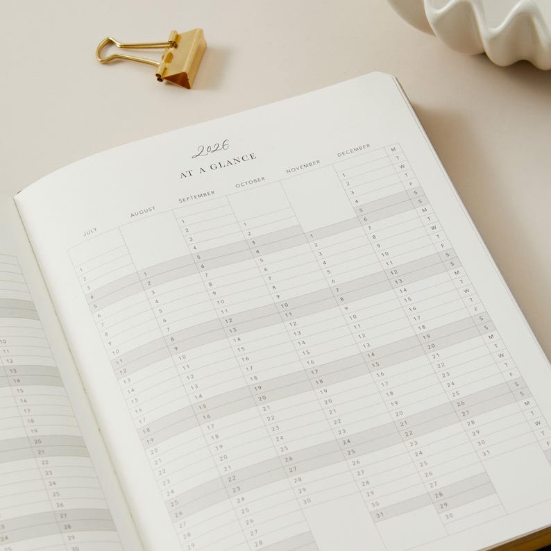 Peut inclure: Agenda ouvert avec un calendrier 2026, pr&eacute;sentant des grilles mensuelles et des dates. Un trombone dor&eacute; est pos&eacute; en haut. Les pages de l'agenda sont claires, avec un design simple et &eacute;l&eacute;gant.