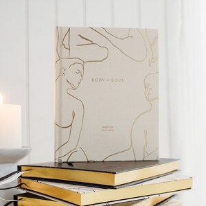 May include: A beige wellness planner with gold line art figures and the words "BODY + SOUL" and "wellness PLANNER". The planner is stacked on top of other books with gold edges.