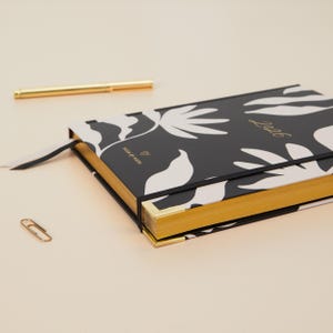 May include: A black and white floral patterned planner with gold gilded edges and the year "2026" in gold lettering. A gold pen, a paperclip, and a black ribbon bookmark are also visible.