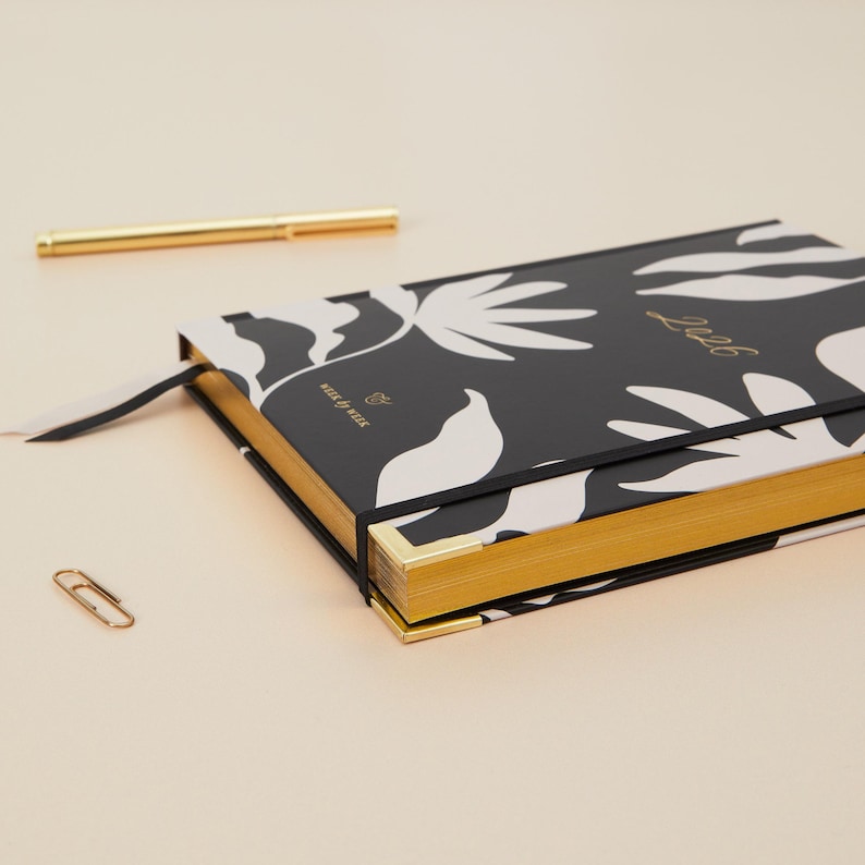 May include: A black and white floral patterned planner with gold gilded edges and the year "2026" in gold lettering. A gold pen, a paperclip, and a black ribbon bookmark are also visible.